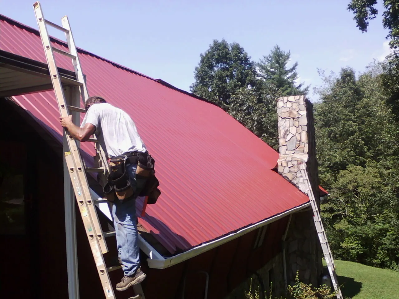 Roofer on a ladder beside a red metal roof and stone chimney — Soffit & Fascia Repair by Roofing Brothers in Slade